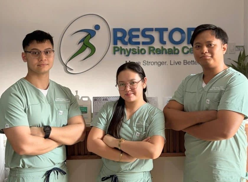 Justin Thettath, Erica Ann Laus, and Zhydie Cantor in clinic scrubs, arms crossed, in front of the Restore Physio Rehab Center sign